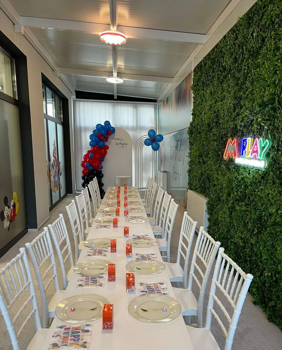The long birthday table set in front of the plant wall and the M Play sign